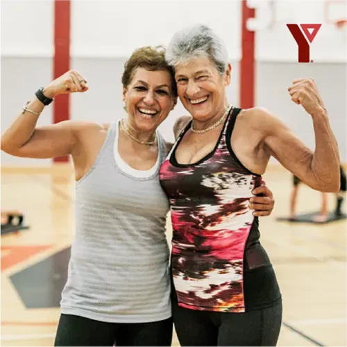 Two elderly women in a gym flexing their biceps and smiling at camera. YMCA logo in top right corner.