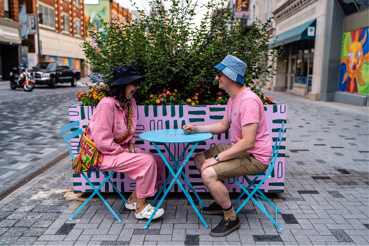 Two citizens dressed in pink, sitting on patio furniture in Downtown London, in front of colourful printed green and pink planter.