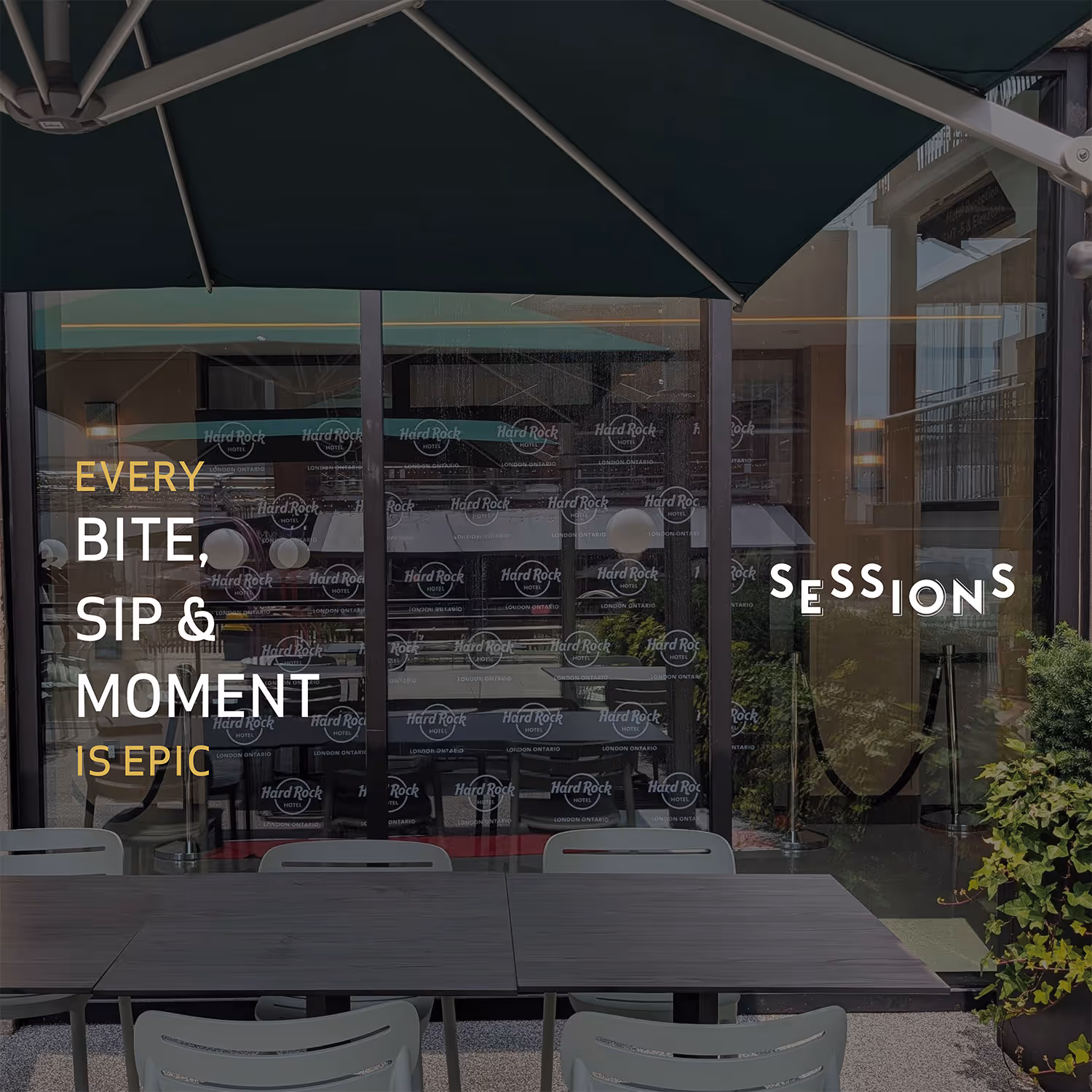 Outdoor seating area with tables and chairs under a green umbrella in front of glass doors featuring Hard Rock Hotel branding and the phrase 'Every bite, sip & moment is epic' and 'Sessions.'