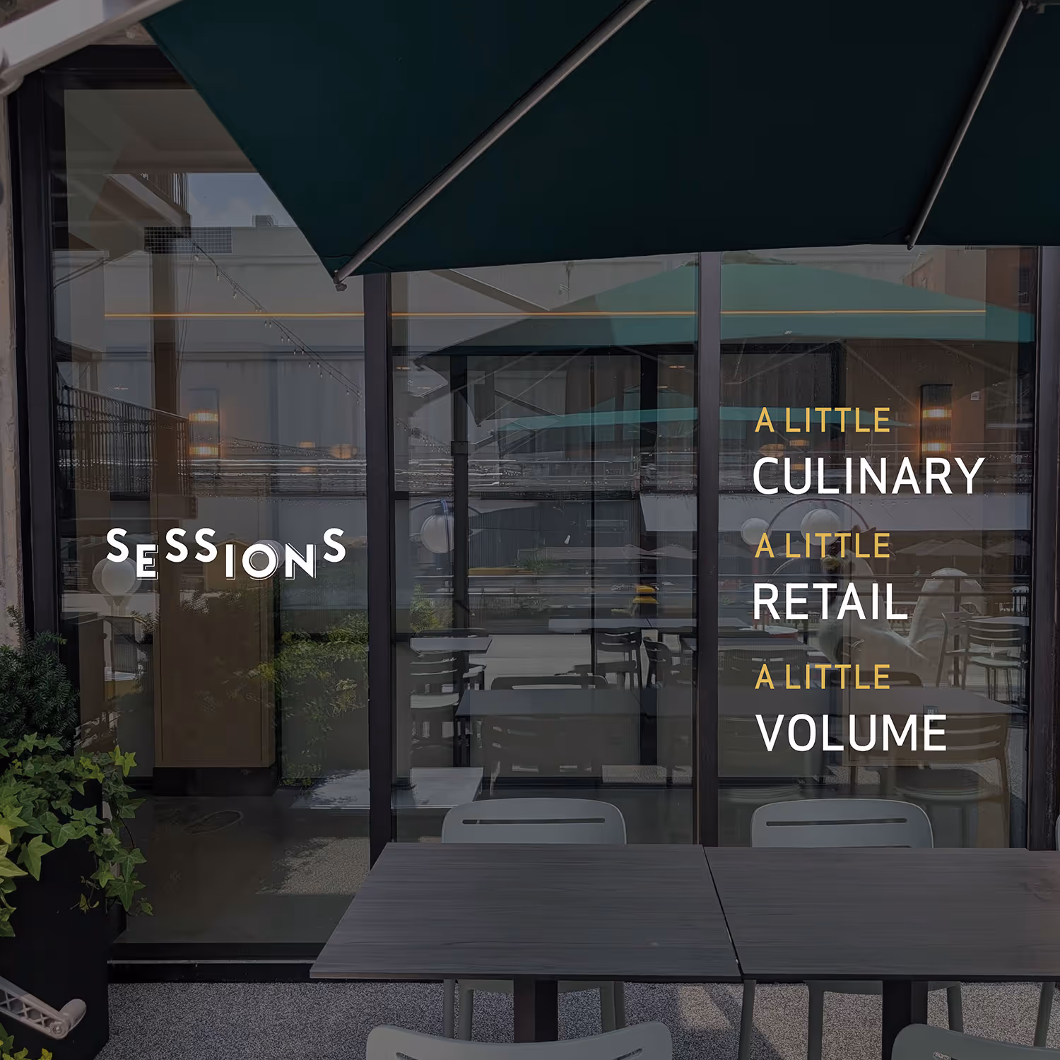 Outdoor seating area with tables and chairs under green umbrella in front of glass windows displaying the words 'Sessions' and 'A little culinary A little retail A little volume'.