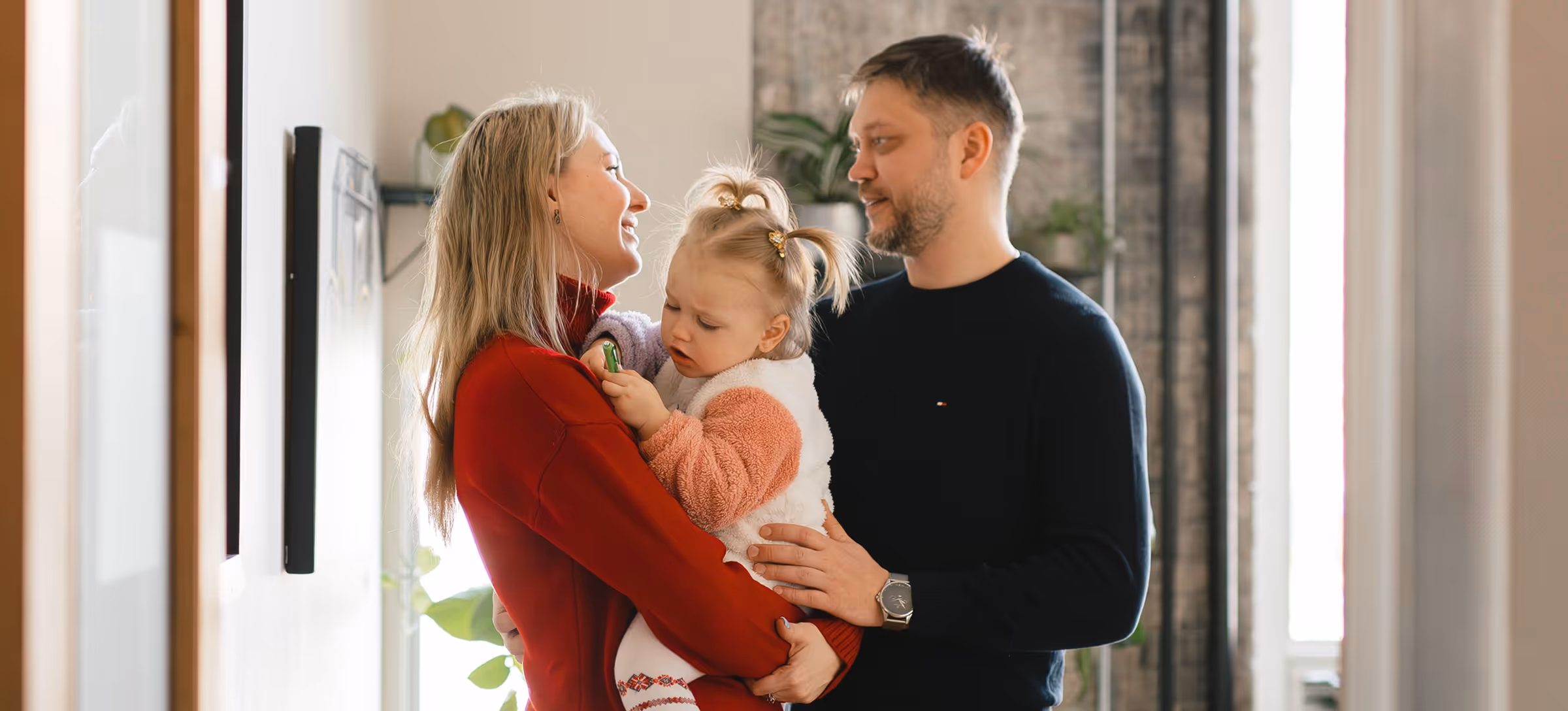 A woman smiling holding a toddler with her husband standing close smiling at them both.