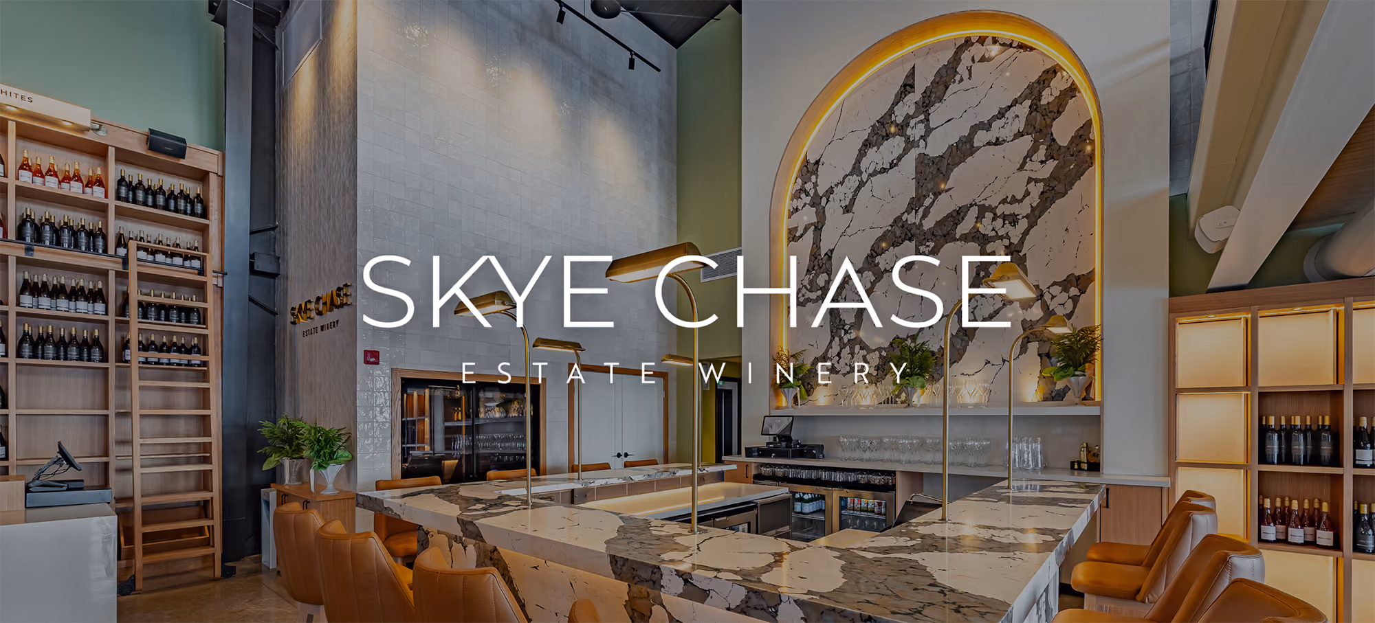 Interior of Skye Chase Estate Winery tasting room featuring a marble-topped bar with seating, modern pendant lighting, wooden shelves with wine bottles, and a decorative marble wall with an arched top.