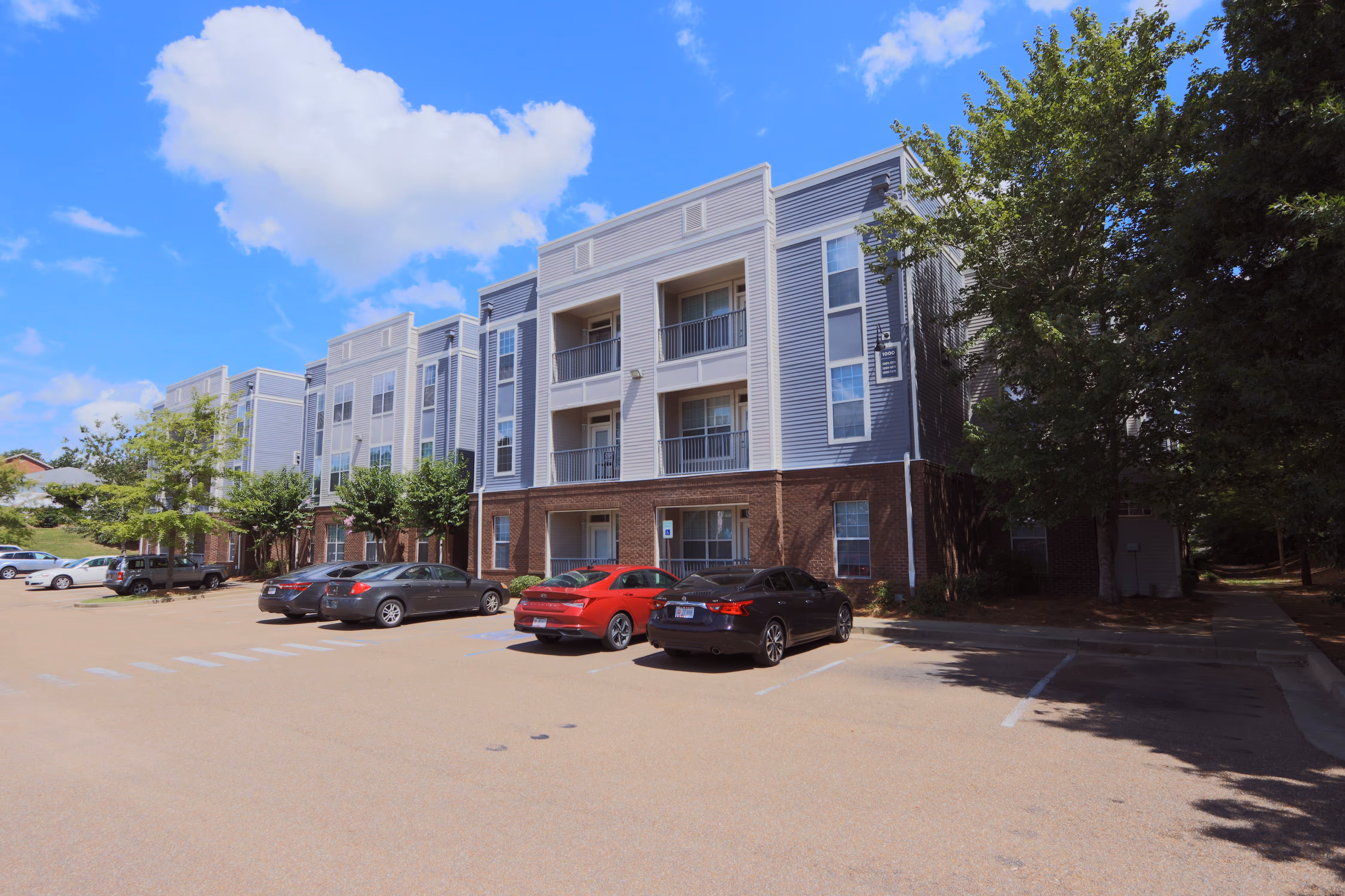 Exterior of the modern apartment building with blue and gray siding, featuring brick accents, balconies, and resident parking at Rev Oxford in Oxford, MS.