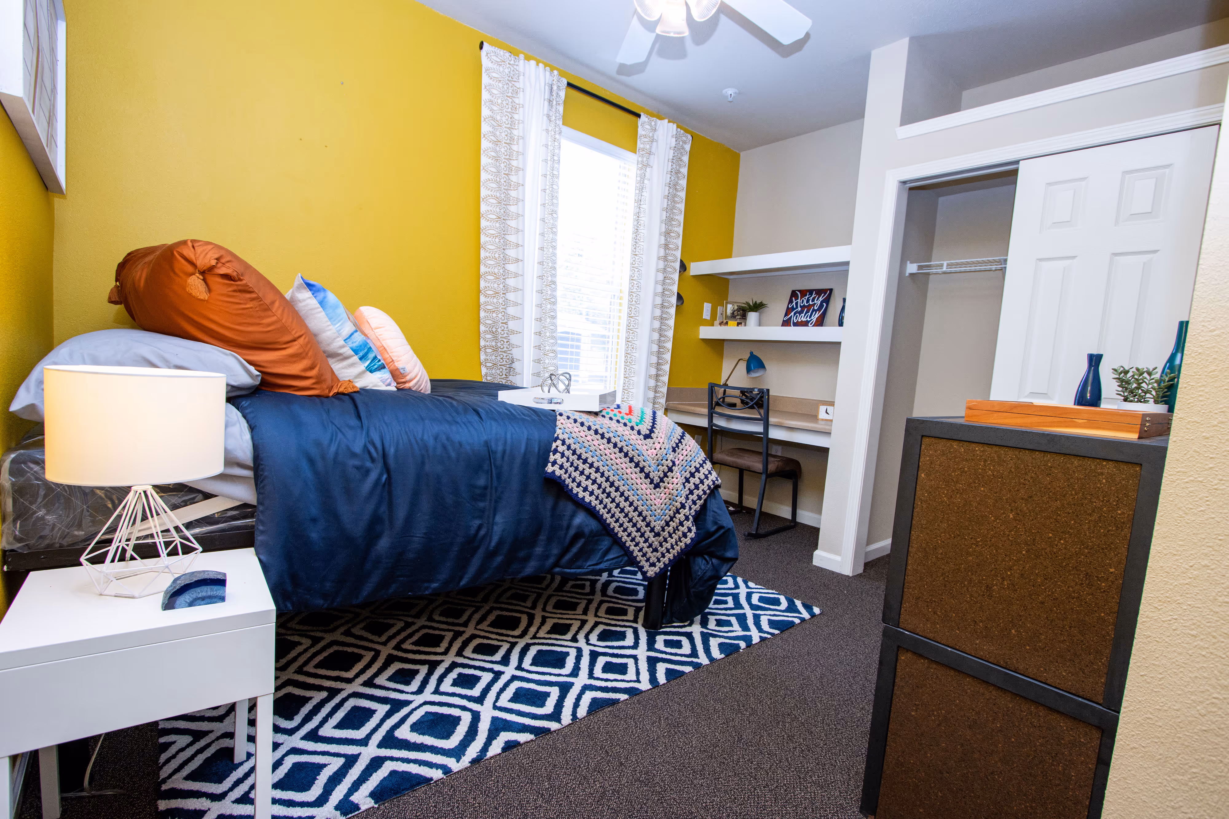 Student apartment bedroom with a bed, desk, and built-in shelving, featuring a vibrant yellow accent wall at Rev Oxford in Oxford, MS.