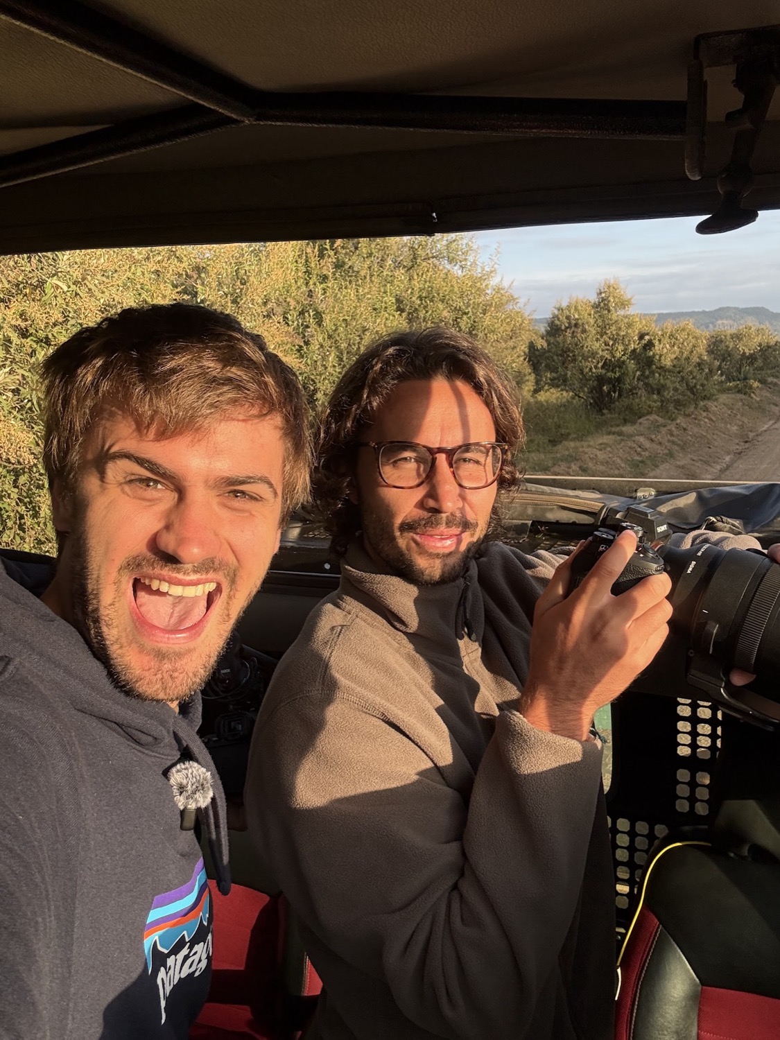 Dos hombres sonrientes en un vehículo de safari, uno sostiene una cámara profesional con teleobjetivo