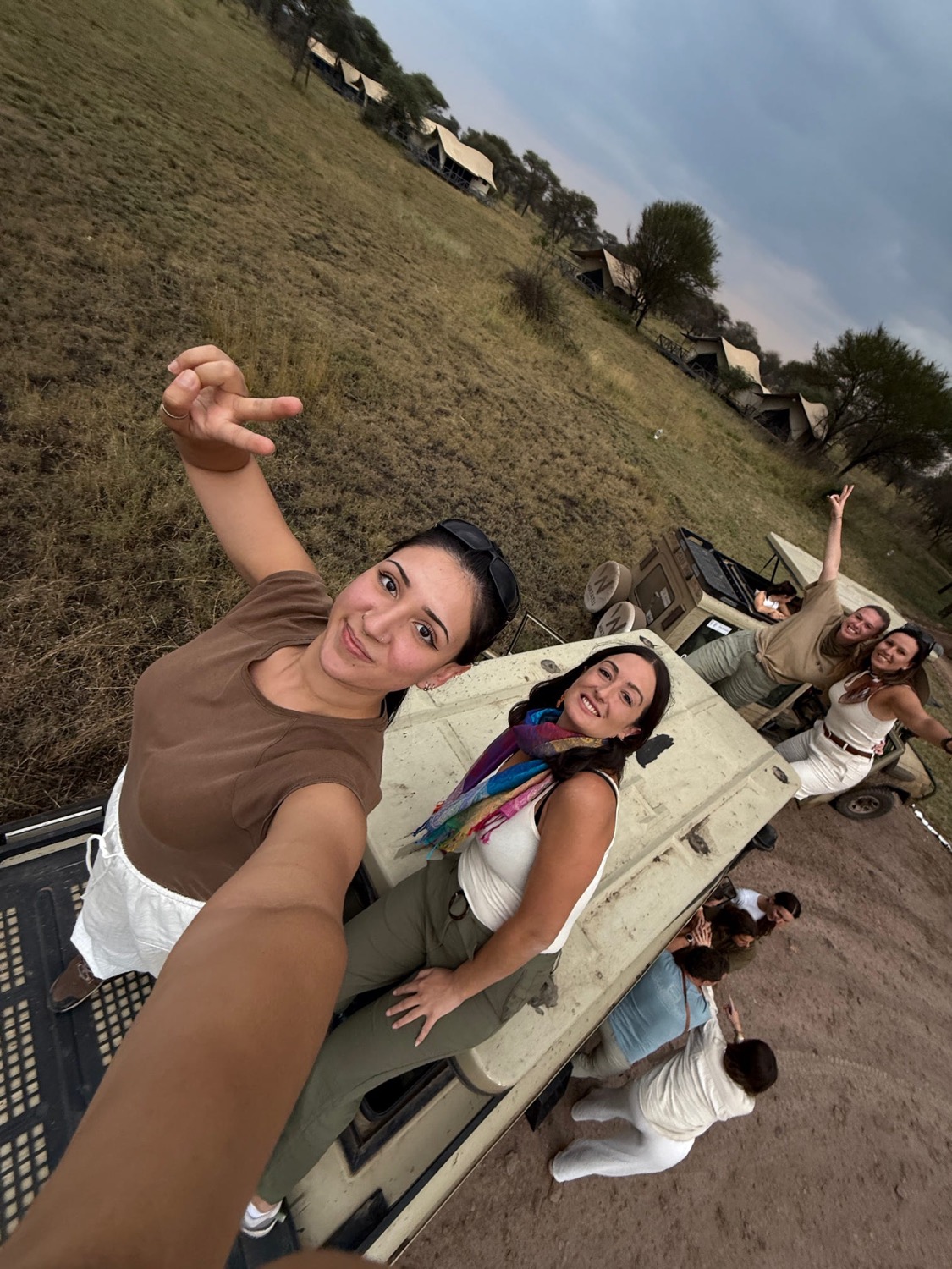 Grup de dones posant alegres vora vehicles de safari amb campament de tendes al fons