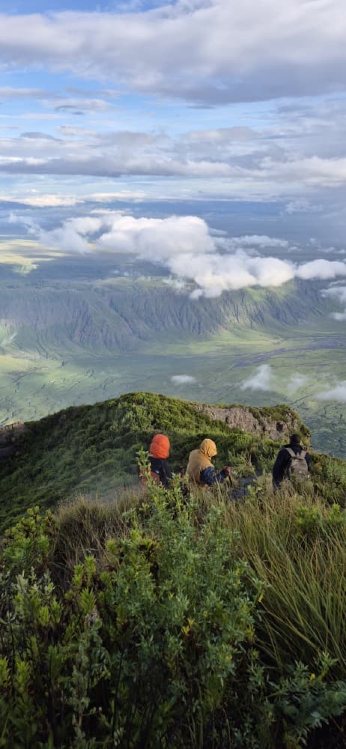 Three backpack-wearing hikers sit on a green mountain ridge overlooking a vast cloud-draped valley below