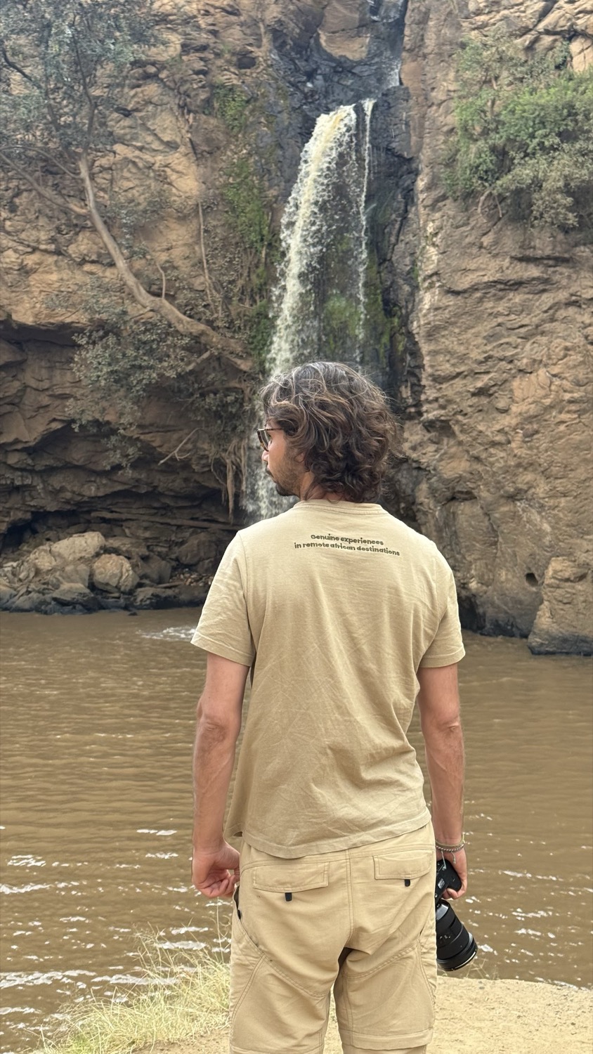 Fotògraf d'esquena contempla una cascada que cau entre roques sobre un riu marró