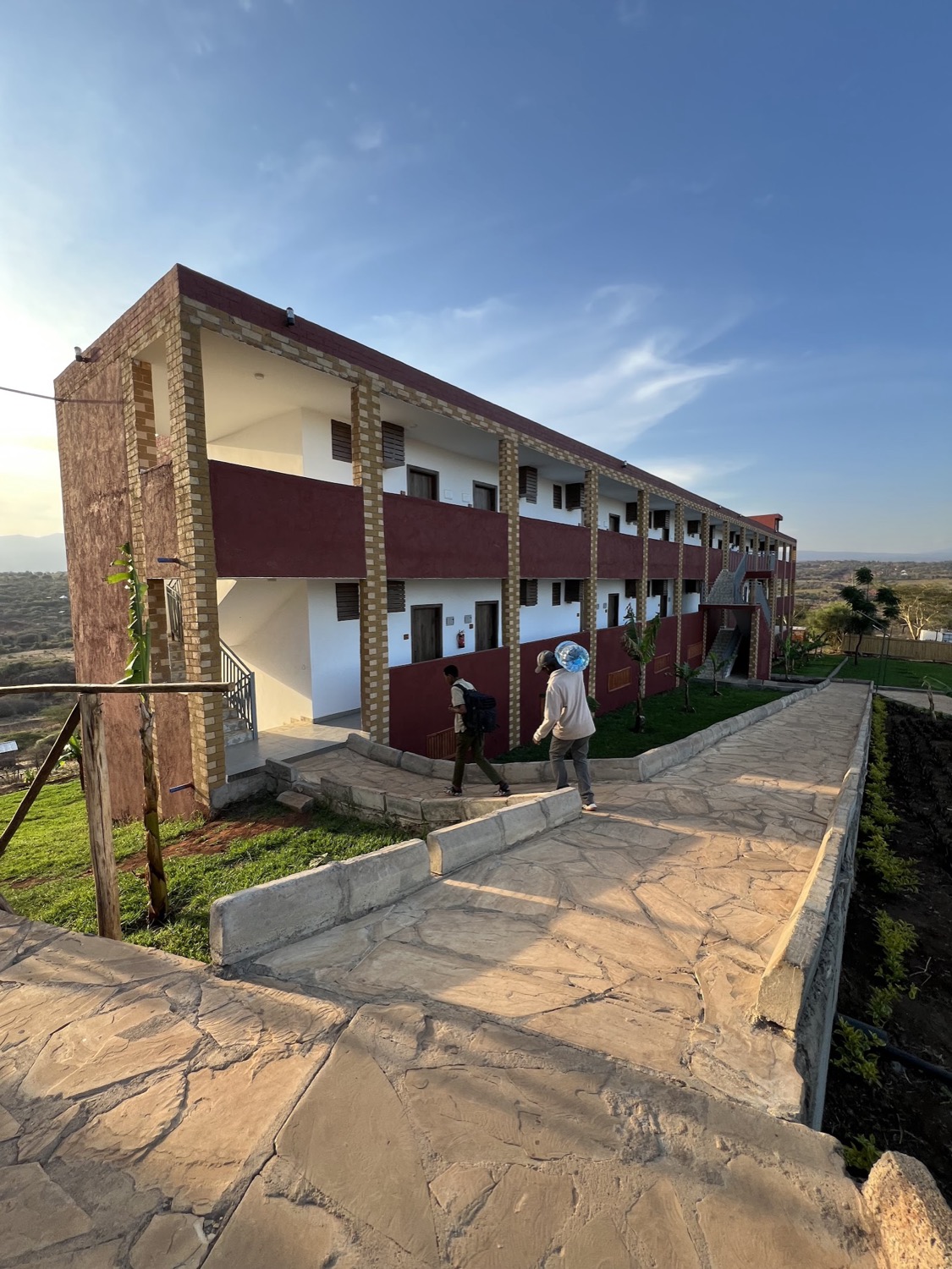 Two people walk along a stone path in front of a two-storey lodge building overlooking a wide African landscape