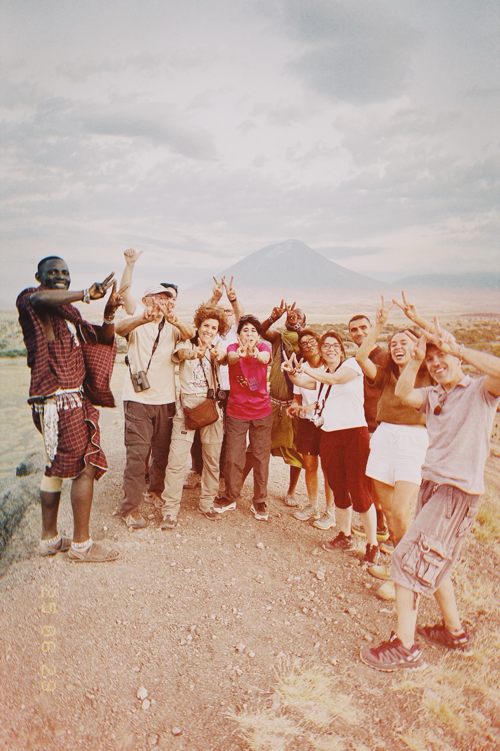 Group of tourists and a Maasai guide smiling and making victory signs before a volcanic cone and arid plain