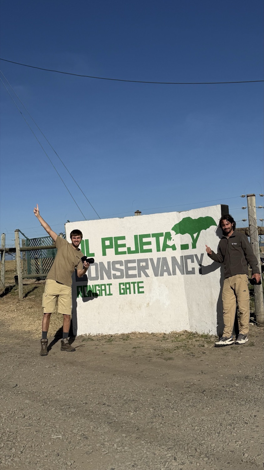 Dos guías posan con entusiasmo junto al cartel de Ol Pejeta Conservancy bajo un cielo azul