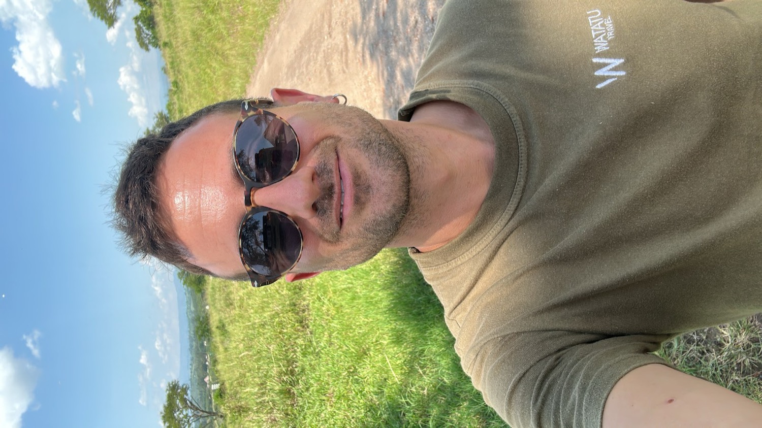 Man wearing sunglasses and a Watatu Travel shirt smiles against a green savanna backdrop