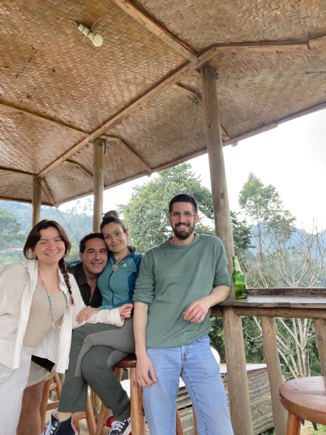 Four smiling people relaxing under a rustic thatched shelter overlooking forested mountains