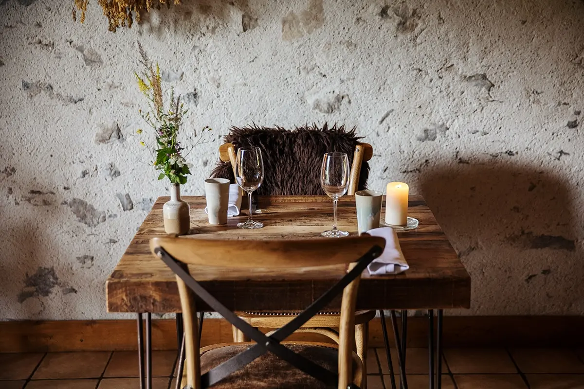 A cozy, rustic table for two is set with wine glasses, candles, and simple decor against a stone wall.