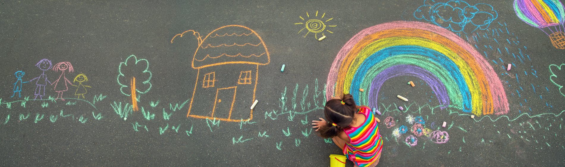 A child in a rainbow shirt drawing a colorful chalk rainbow, sun, and house on the pavement