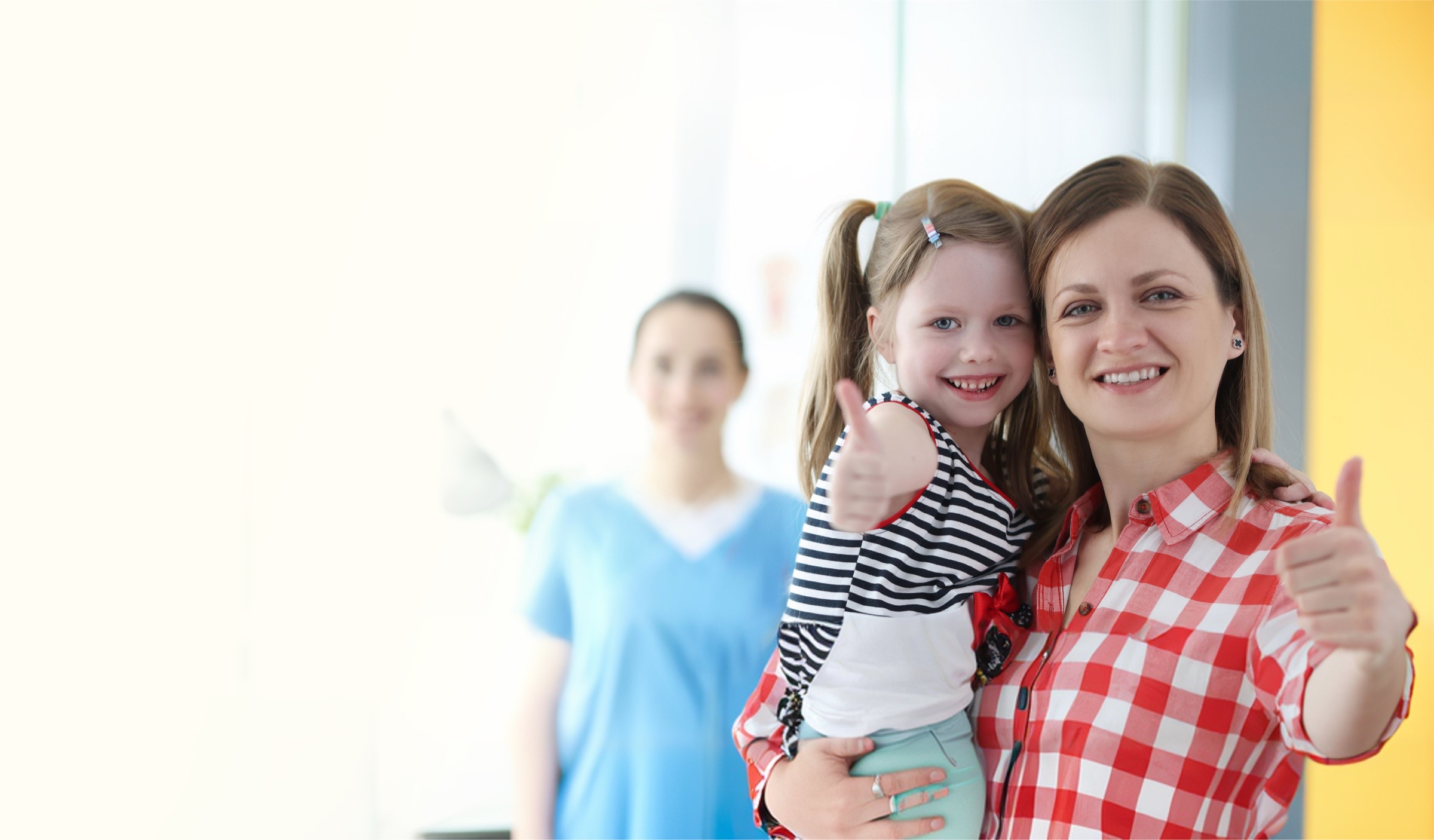 A smiling mother holding her young daughter giving a thumbs-up, with a clinician in the background