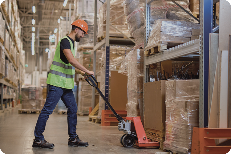 Man in safety gear pushes a red pallet jack through a bright, organized warehouse filled with tall shelves and wrapped boxes on pallets.
