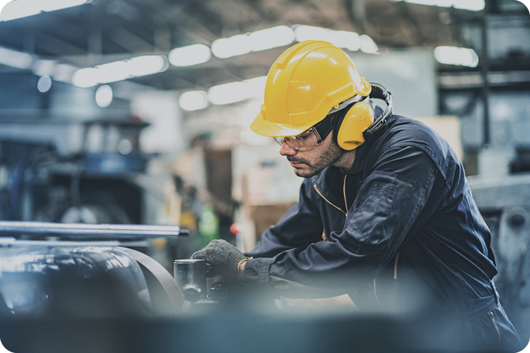 Medium shot of a man in navy coveralls, yellow hard hat, and gloves operating machinery in a large, blurred industrial or manufacturing space.