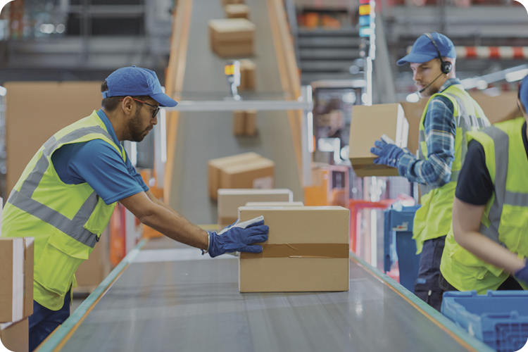 Full warehouse shot with three workers in safety vests handling boxes along a conveyor belt, surrounded by equipment in a busy, well-lit space.