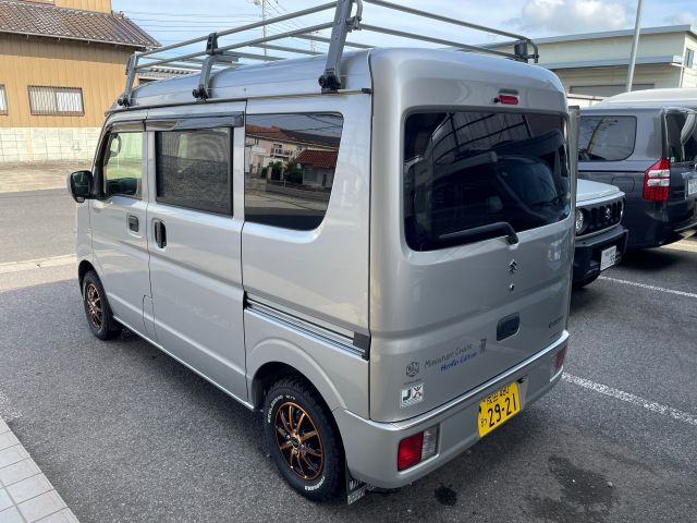 Silver compact van parked on street with roof rack and small bronze-colored rims, viewed from rear left side in a residential area.
