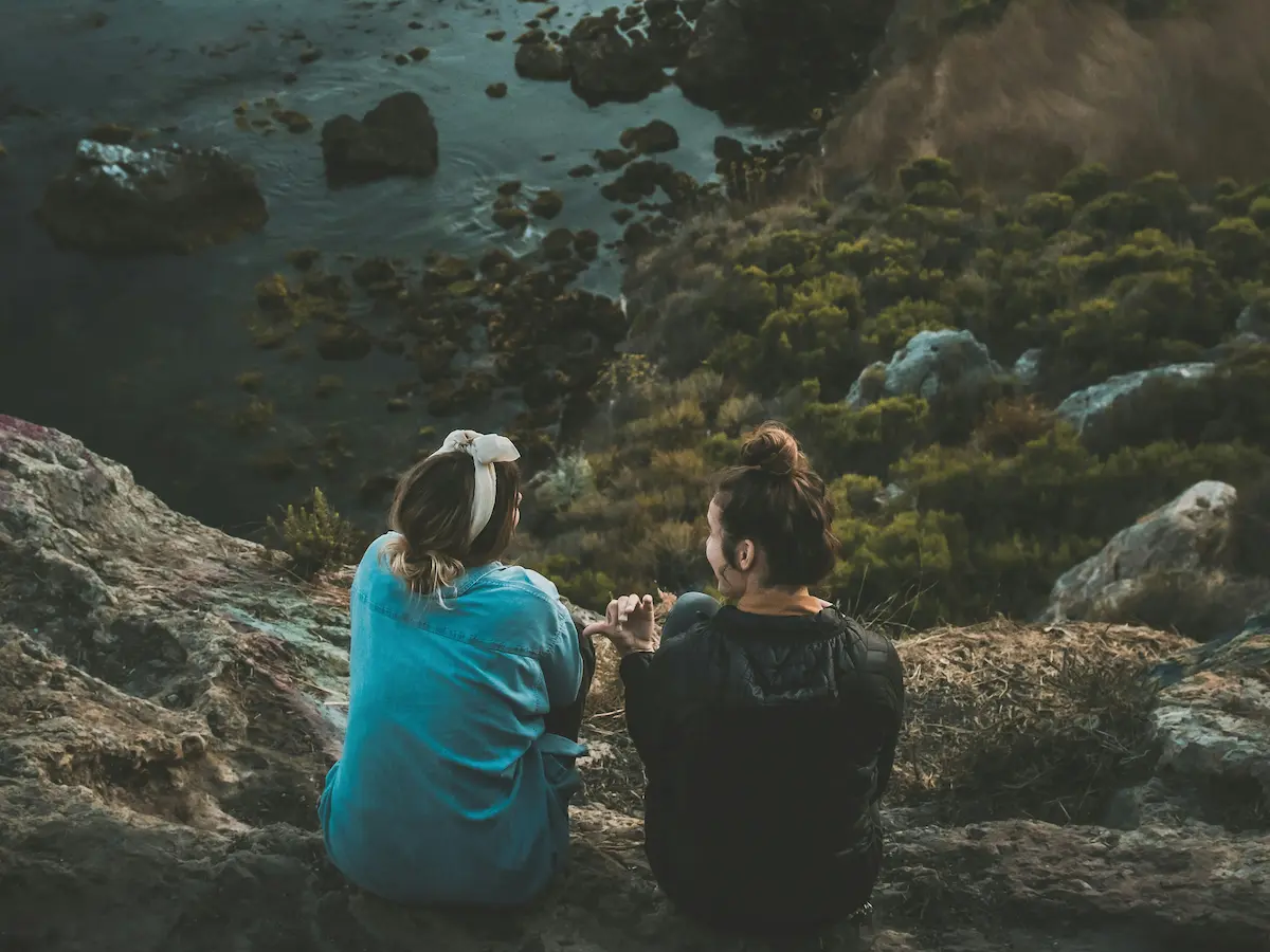 Two people sit by a rocky beach inlet, having a discussion symbolic of someone offering support to another who could be struggling with depressioin.