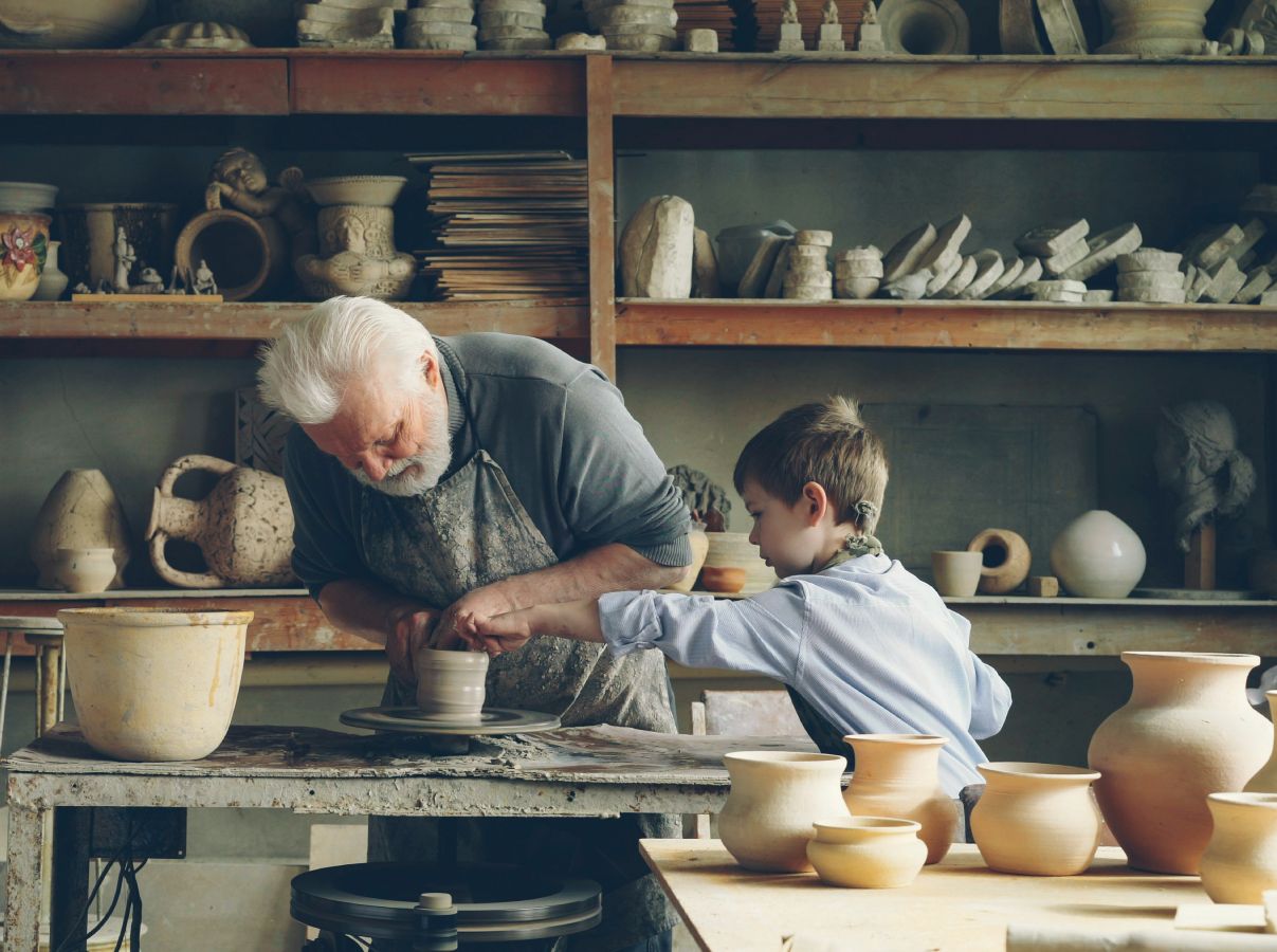 An older person and child work a pottery wheel together as symbolism of senior independence being supported through creativity and sharing knowledge .