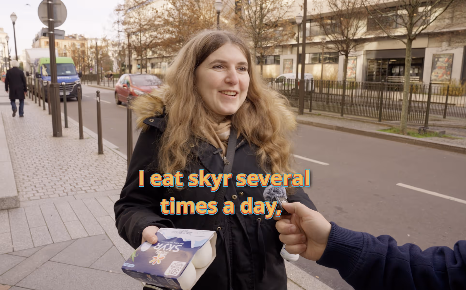 Une jeune femme souriante en manteau noir tenant une boîte de skyr lors d'une interview en rue, un micro pointé vers elle.