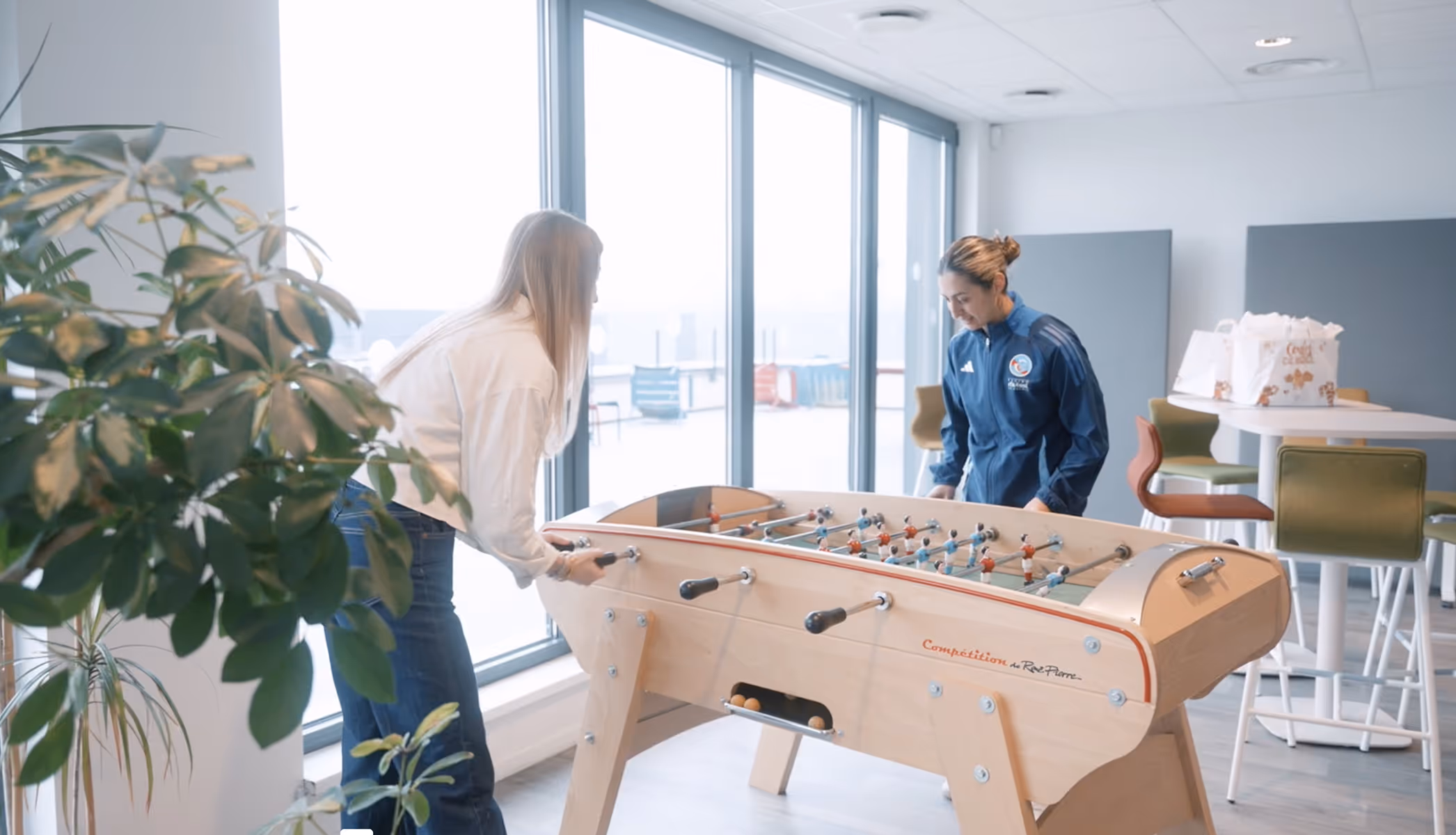 Deux personnes jouent au baby-foot dans une salle lumineuse avec une grande fenêtre et des plantes.