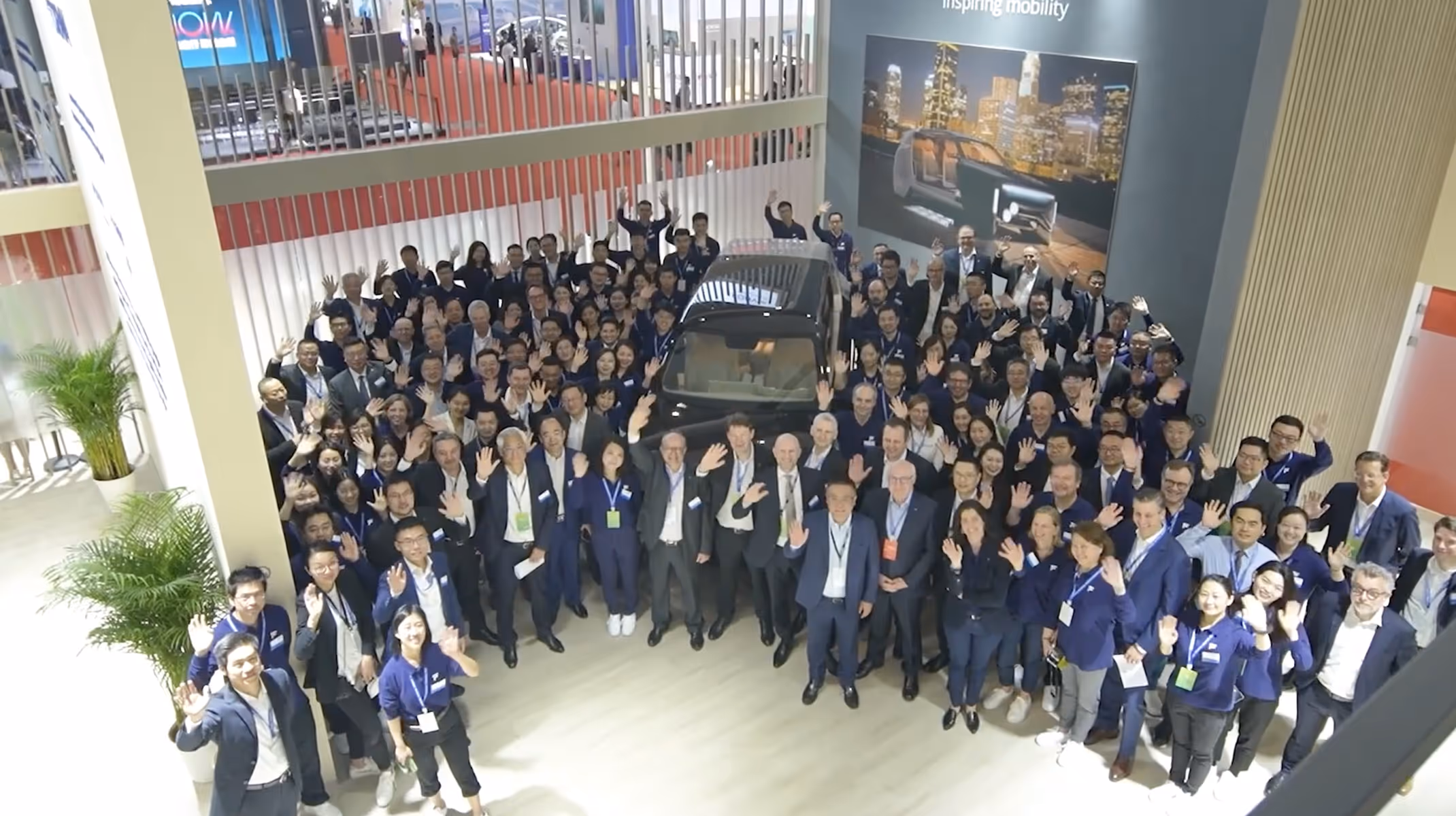 Groupe de nombreuses personnes souriantes posant autour d'une voiture exposée lors d'un événement en intérieur.