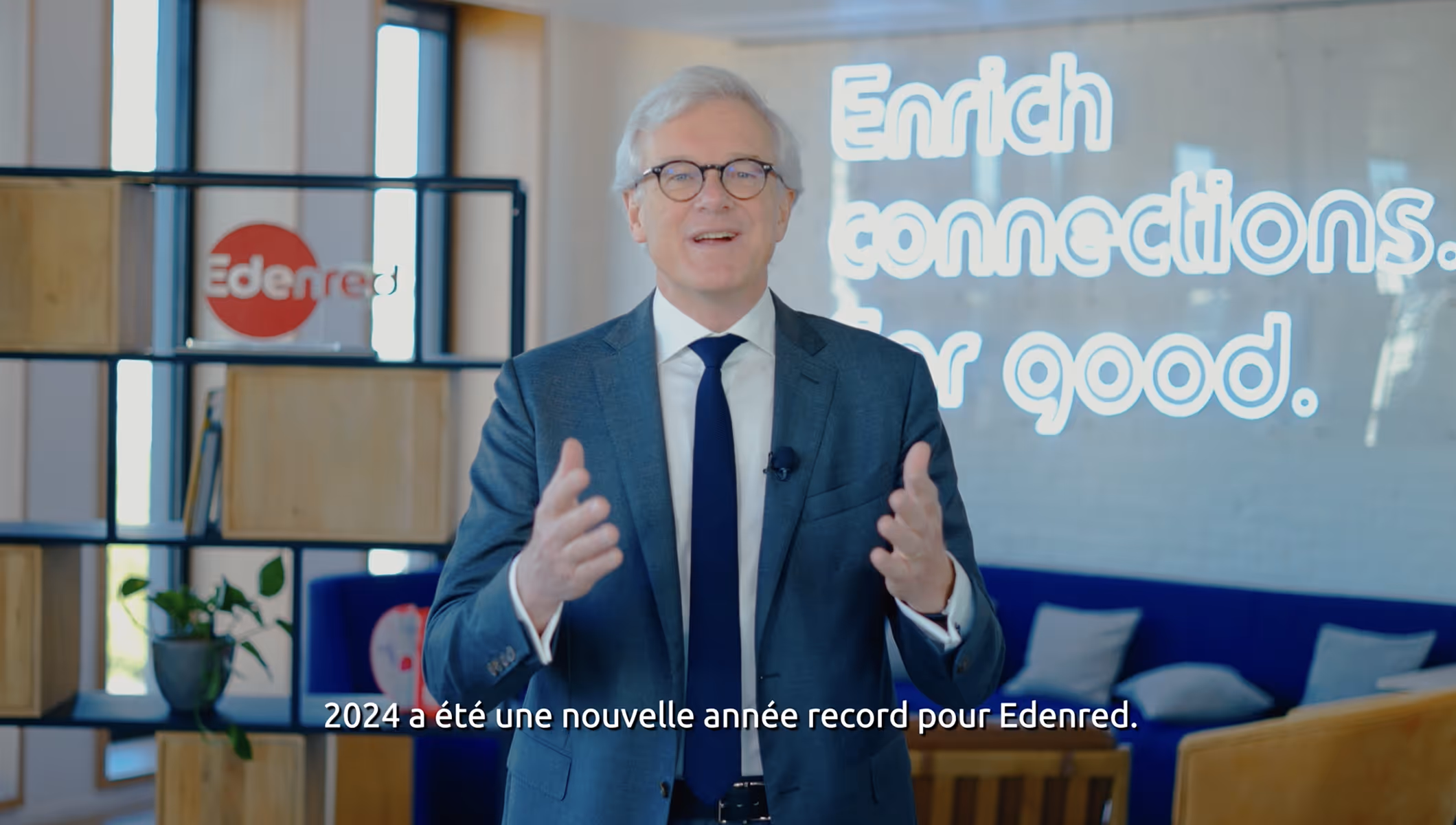 Homme en costume et lunettes parlant devant un écran avec le texte 'Enrich connections. For good.' dans un bureau moderne.