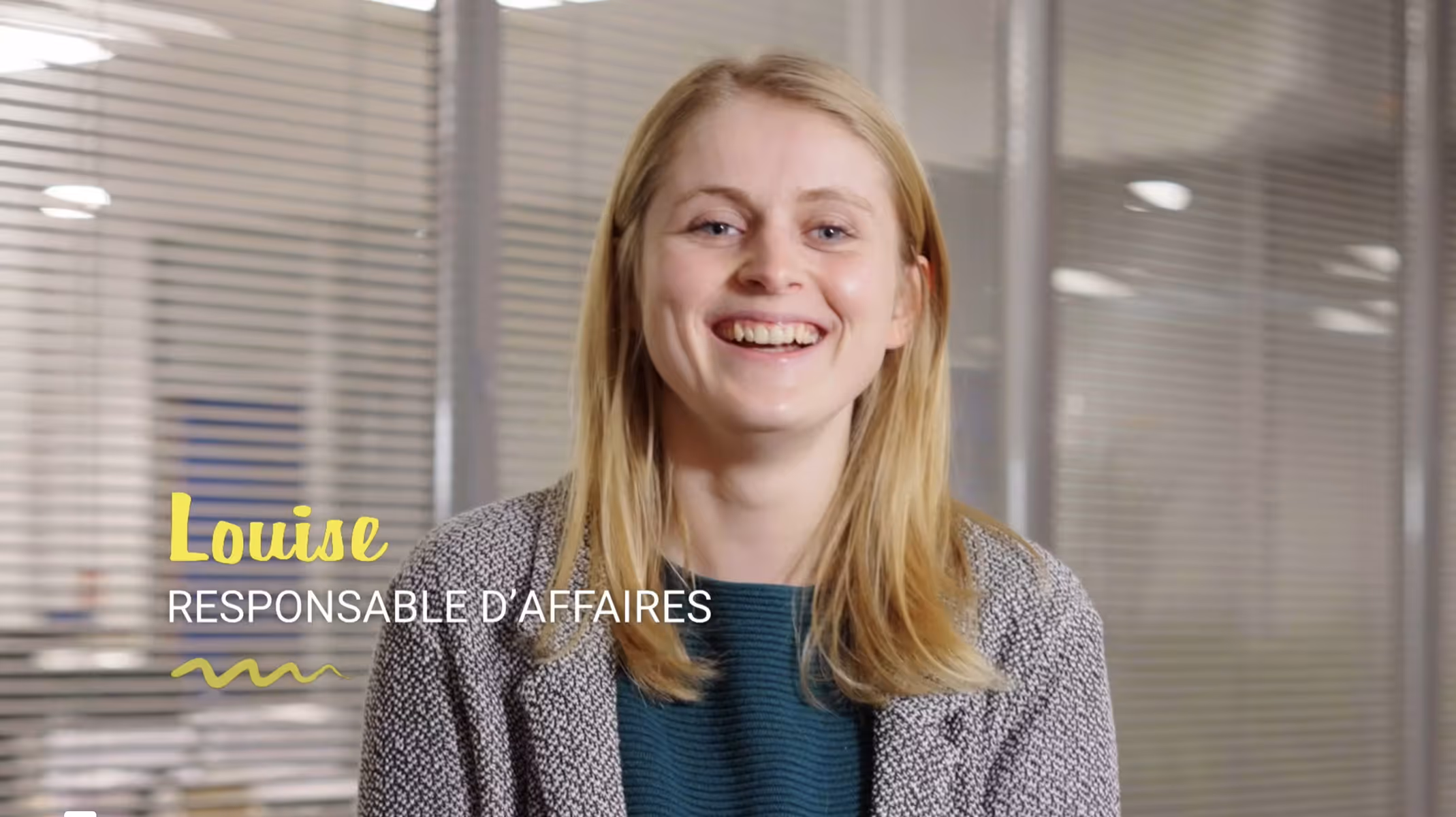 Portrait de Louise, responsable d'affaires souriante, devant une cloison en verre avec stores.
