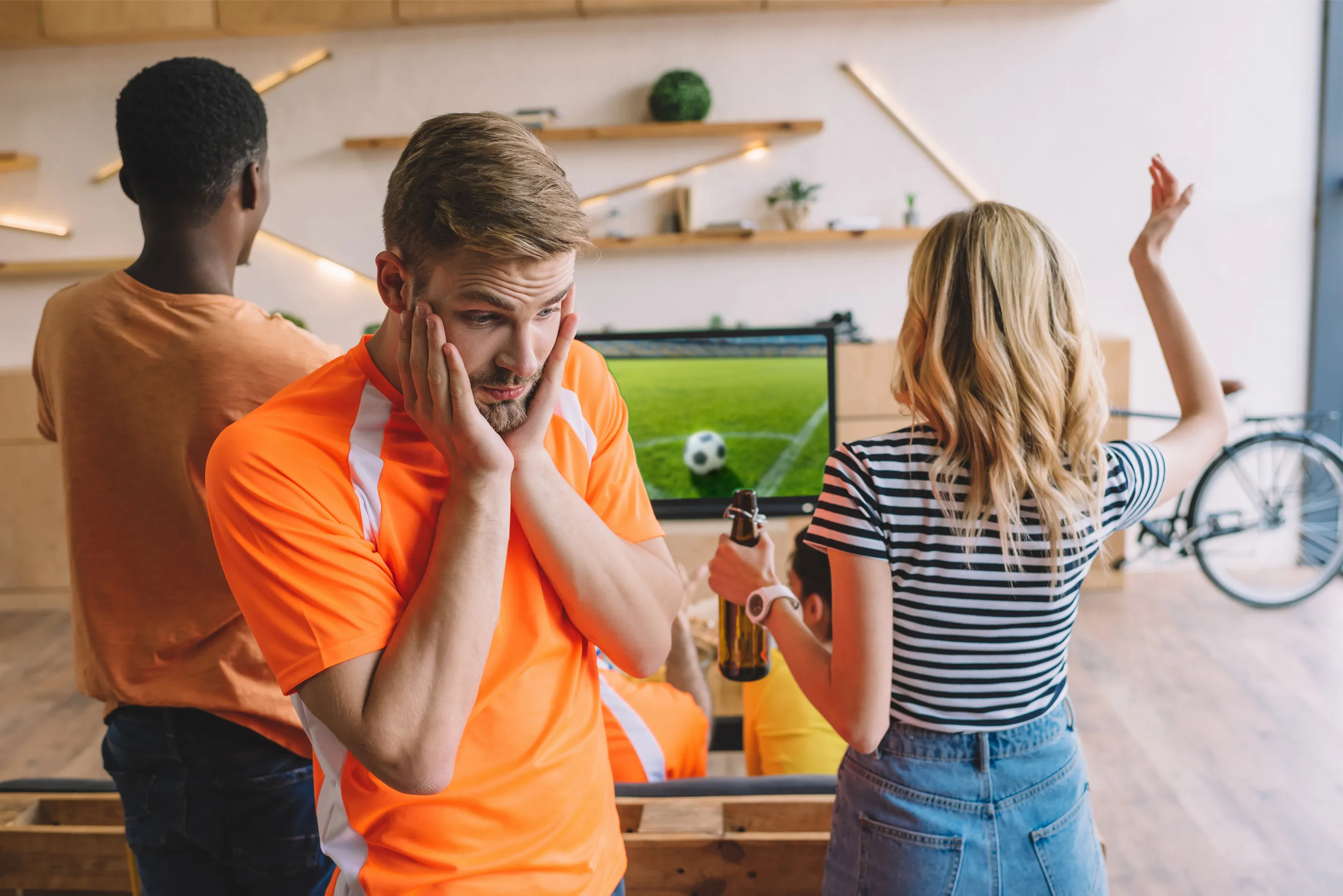 Distressed soccer fan in a bright orange jersey holds his face in his hands while watching a game on TV. A woman next to him, wearing a striped shirt, throws her arm up in reaction.