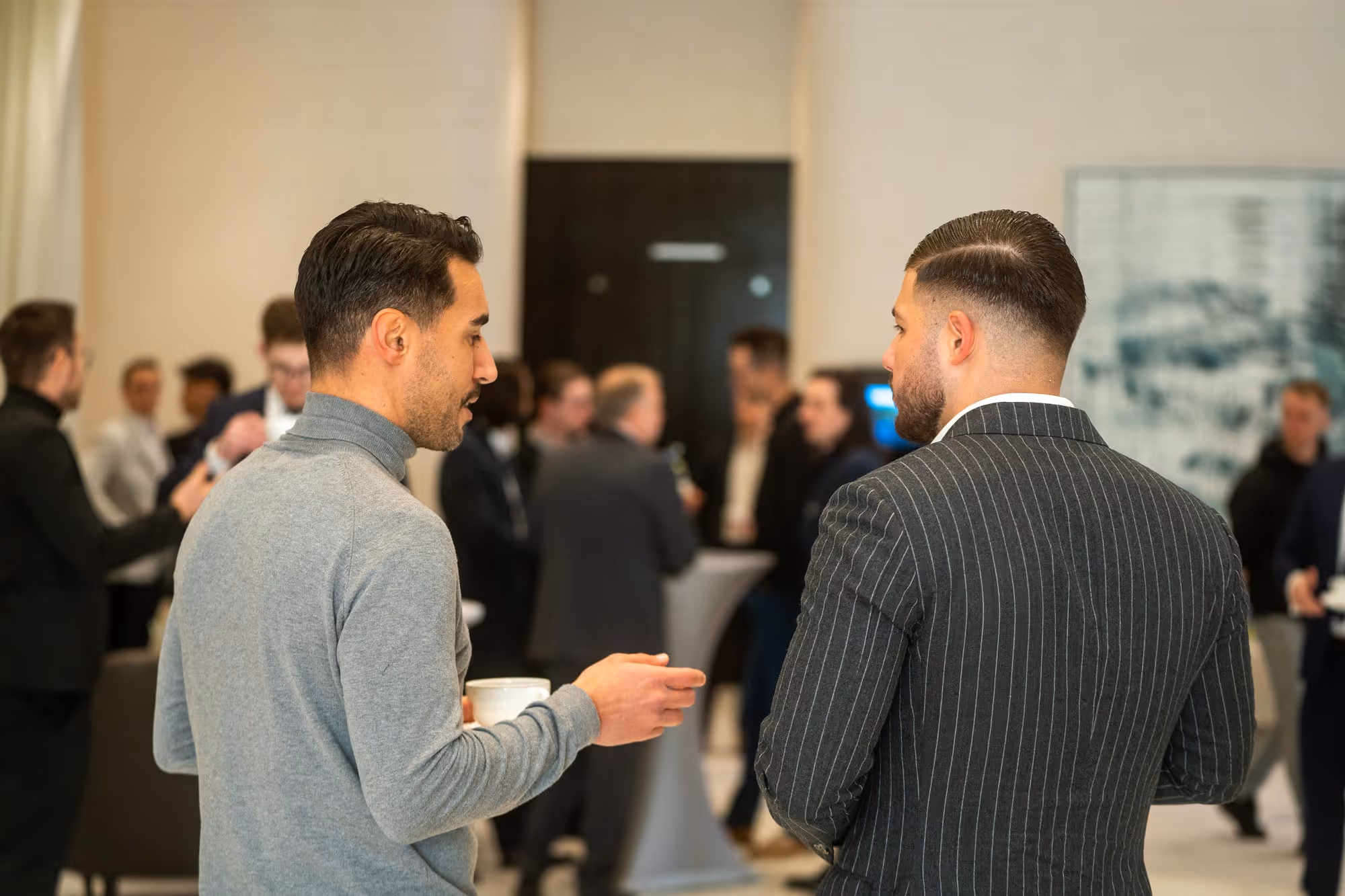 Zwei Männer unterhalten sich bei einem geschäftlichen Empfang, einer hält eine Kaffeetasse; im Hintergrund weitere Gäste.