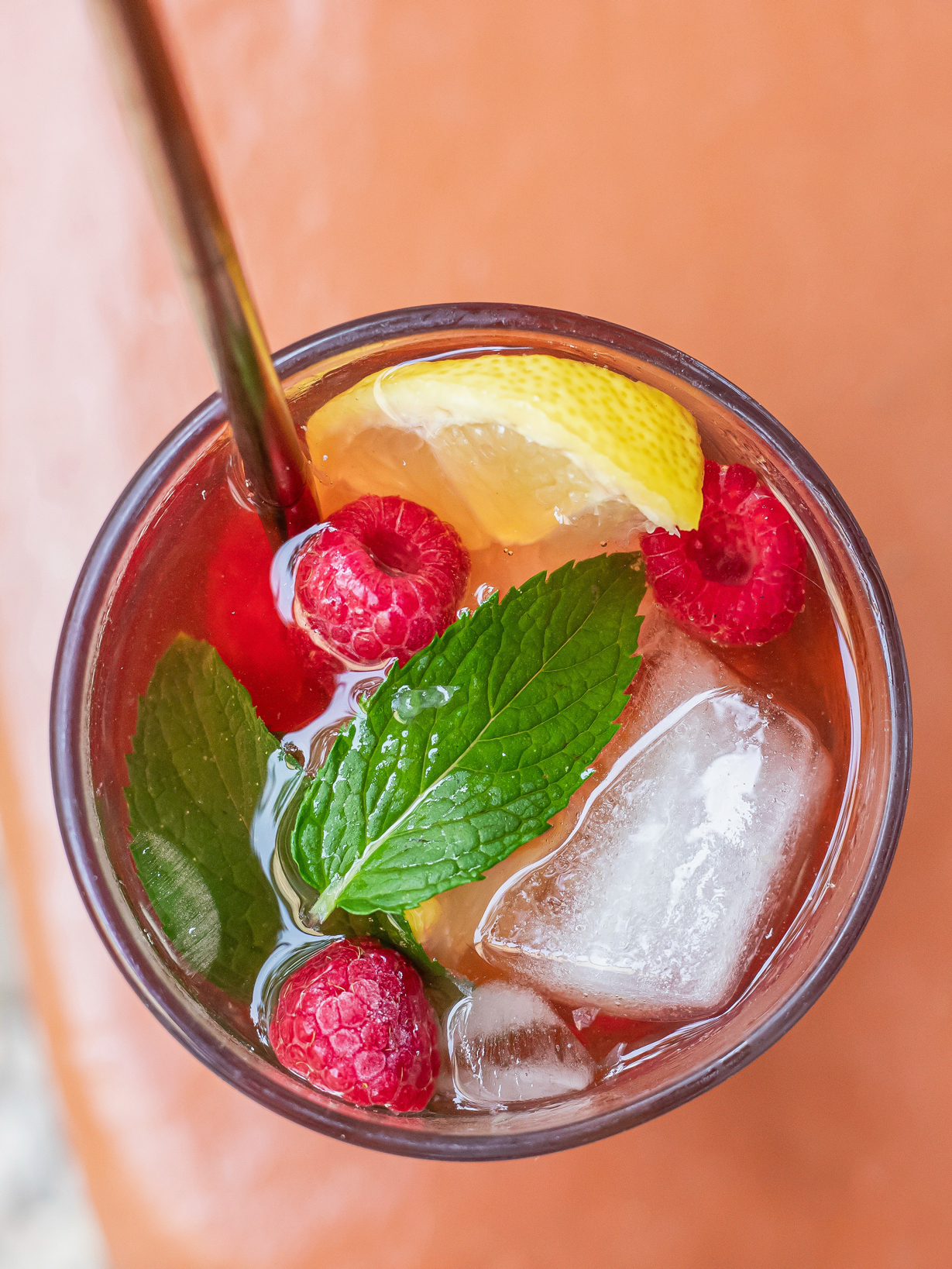 Boisson glacée rafraîchissante servie dans un verre transparent, garnie de glaçons, de framboises et d’une tranche de citron. Le verre est posé sur une table, avec un fond flou évoquant une ambiance estivale et lumineuse.