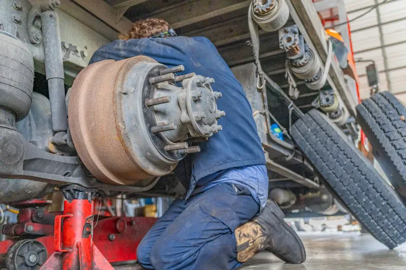 Truck repair technician servicing the brake drum and wheel hub assembly with the tire removed inside a heavy-duty repair shop.
