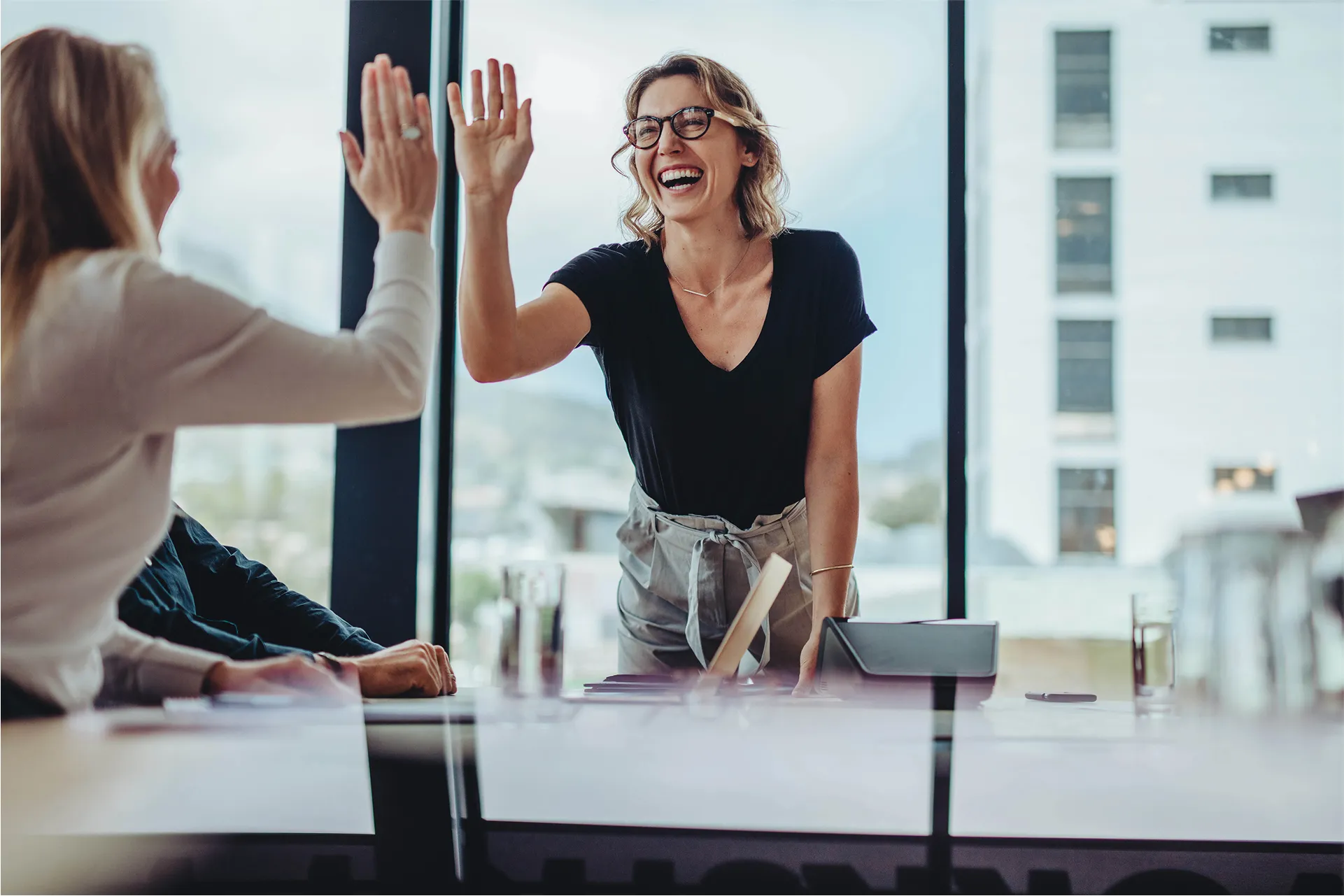 Smiling woman in glasses giving a high-five to a colleague during a positive team meeting in a modern office.