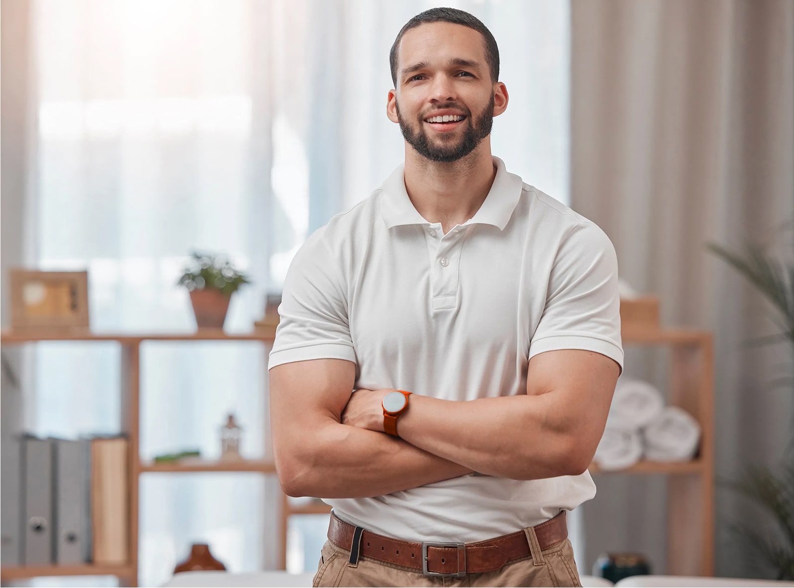 Smiling man in a white polo shirt standing with arms crossed in a modern office or home workspace.
