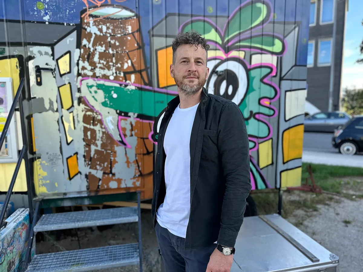 Dr. Daniel Schmelzer standing outdoors in front of a colorful graffiti wall, wearing a black jacket over a white t-shirt, looking slightly to the side.