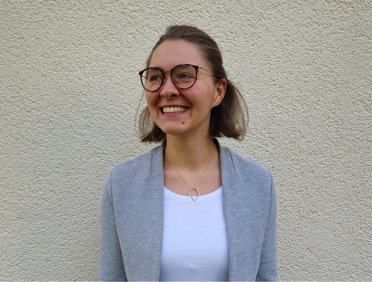 Sabrina Fröhlich smiling, wearing glasses, a light grey blazer and white top, standing against a neutral background.