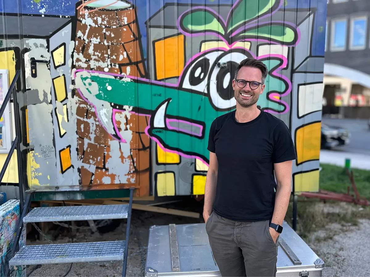 Christian Uetz smiling, wearing glasses, a black t-shirt and grey trousers, standing in front of a colorful graffiti wall