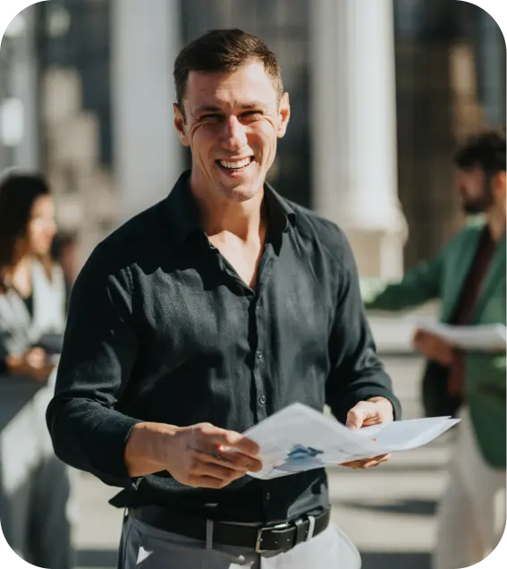 Business professional smiling while holding documents outdoors, with colleagues in the background.