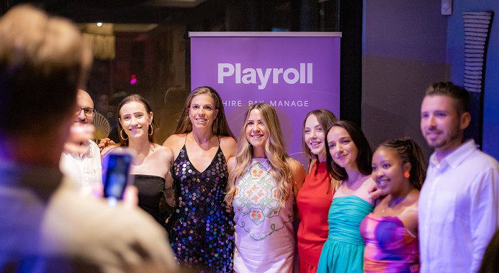Six smiling women posing for a photo at a Playroll event with a purple banner in the background.