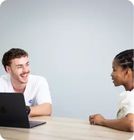 Two people sitting at a table, smiling and talking, with a laptop in front of one person.
