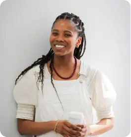 Smiling woman with braided hair wearing a white dress, holding a coffee cup.