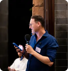 Man with glasses holding microphone and phone speaking at an event against a dark background.