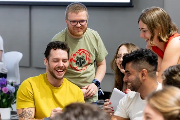 Group of six diverse young adults smiling and looking at a smartphone together indoors.