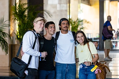 Four young adults smiling and posing for a photo indoors with backpacks and casual clothes.