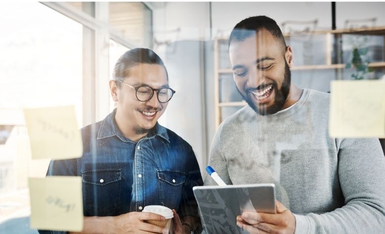 Graphic showing two colleagues smiling while reviewing information on a tablet in an office, representing collaboration and productivity for a workforce management software blog.