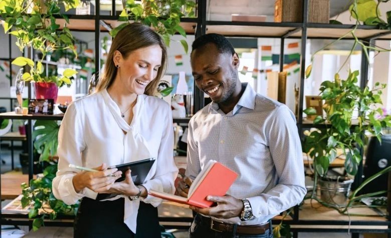 Grafik, die zwei lächelnde Kollegen zeigt, die gemeinsam Notizen in einem modernen Büro durchgehen und symbolisiert, wie offene Gespräche und Feedback das Mitarbeiterengagement fördern und bei der Messung des Employee Net Promoter Score (eNPS) helfen.