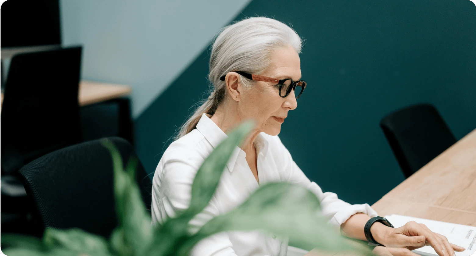 Image of a professional woman focused on her work, with a plant in the foreground in an office setting.